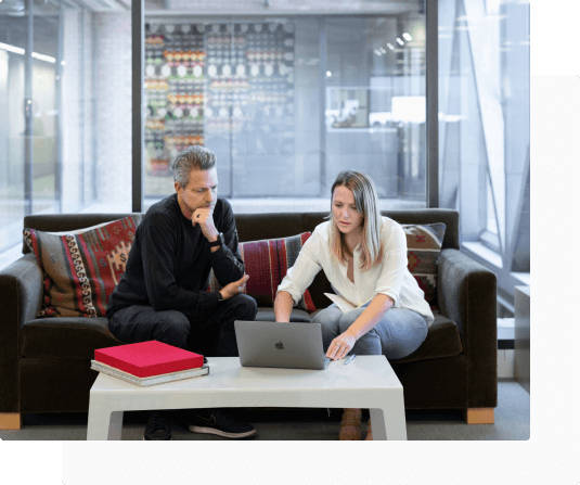 Two colleagues, a male and female, sitting on a coach in a meeting room looking at a laptop